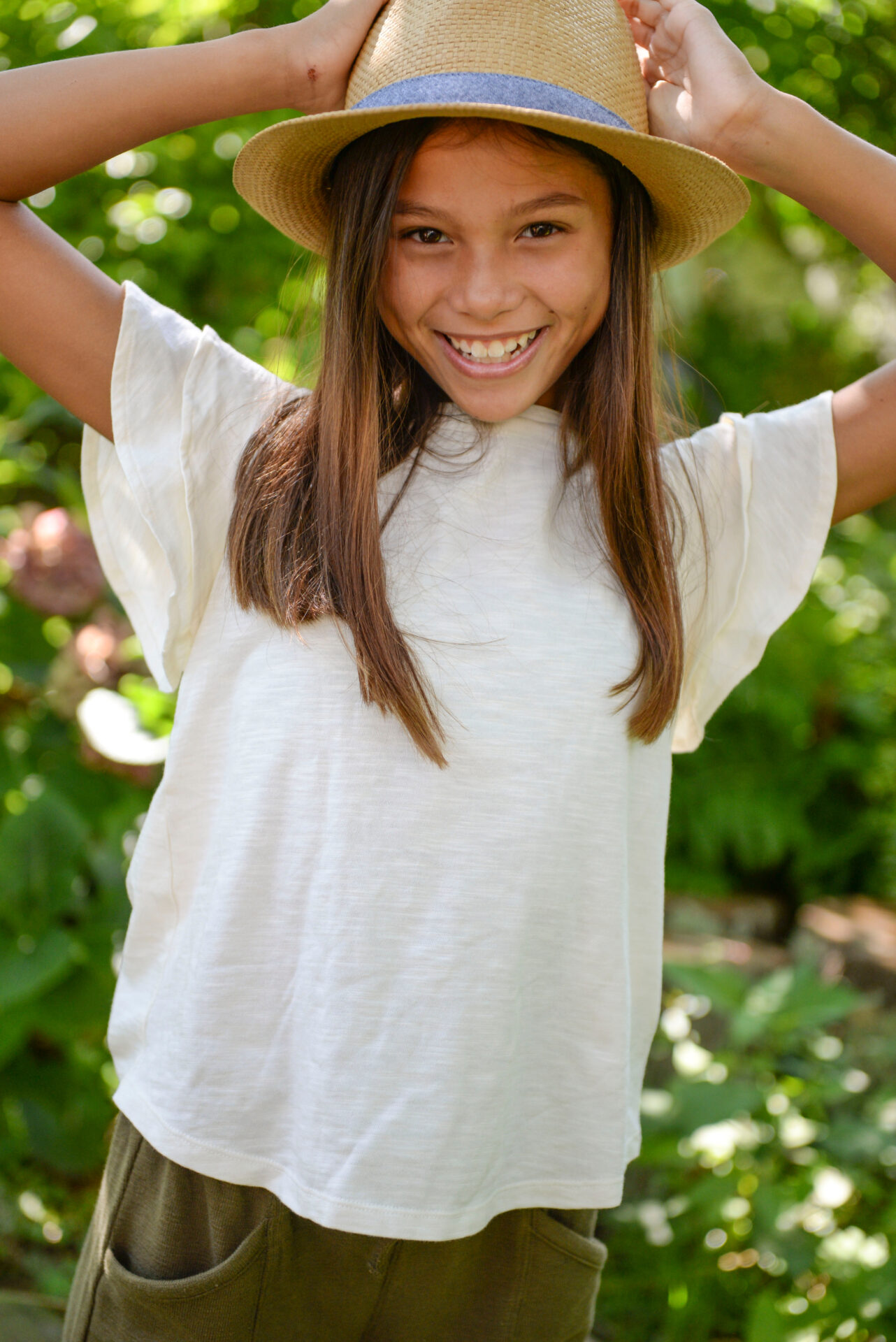A teen smiling at the camera wearing a white top and a woven bucket hat
