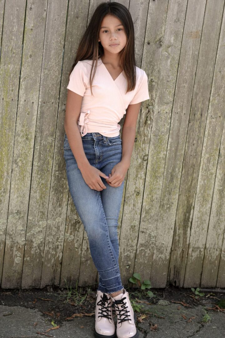 A teen leaning against the wall wearing a pink shirt, denim pants, and pink boots